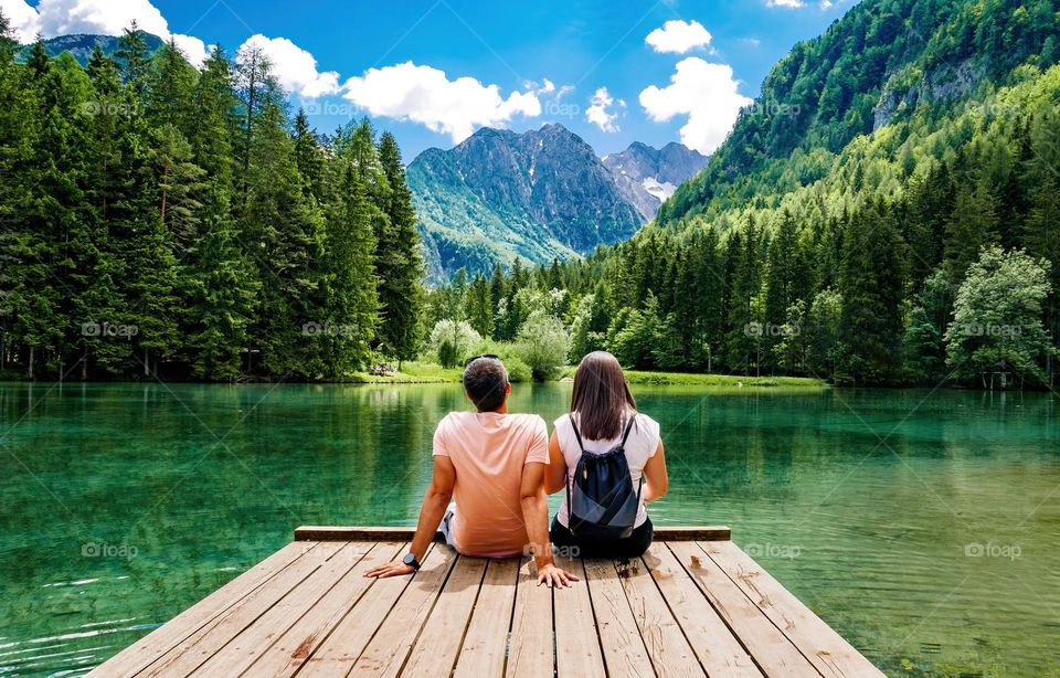 Rear view of young couple sitting on wooden pier on an idyllic lake in mountains on a beautiful sunny day in summer