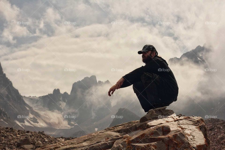 A man in a cap sits on a huge piece of rock in the mountains on a glacier