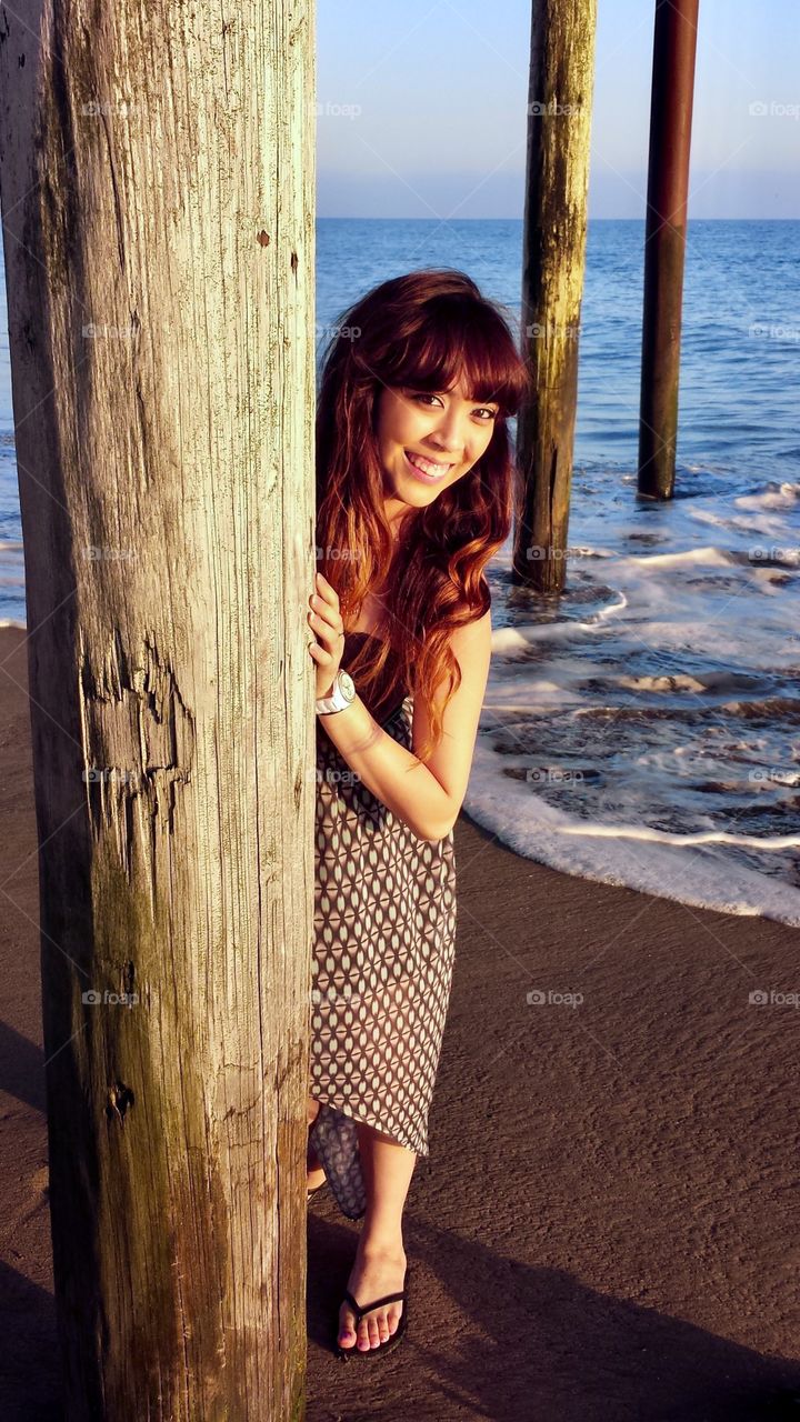 Young woman playfully peeks out from behind a wooden pole under a pier on the beach at low tide