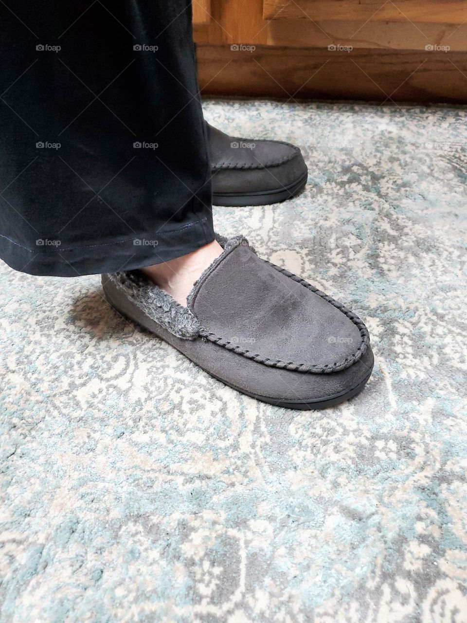 Gray slippers on man standing on rug. Standing on rug keeping feet warm. View of slippers feet on floor in room.