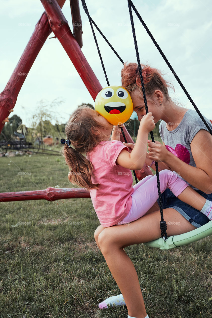 Teenage girl playing with her younger sister in a home playground in a backyard. Happy smiling sisters having fun on a swing together on summer day. Real people, authentic situations