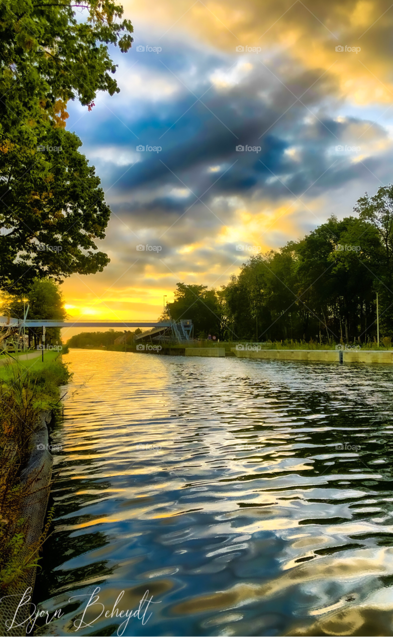 Dark stormy clouds and a golden sunrise combined in the sky over a river or canal, reflected in the water