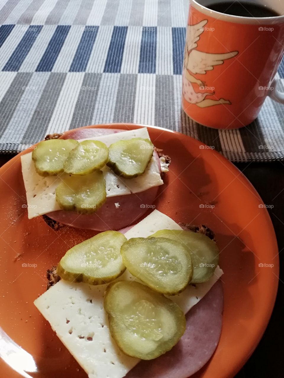 Two slices of dark bread with butter, ham, cheese, pickled cucumber and some crumbs on an orange plate on a brown table. A cup of black coffee on a striped table cloth.