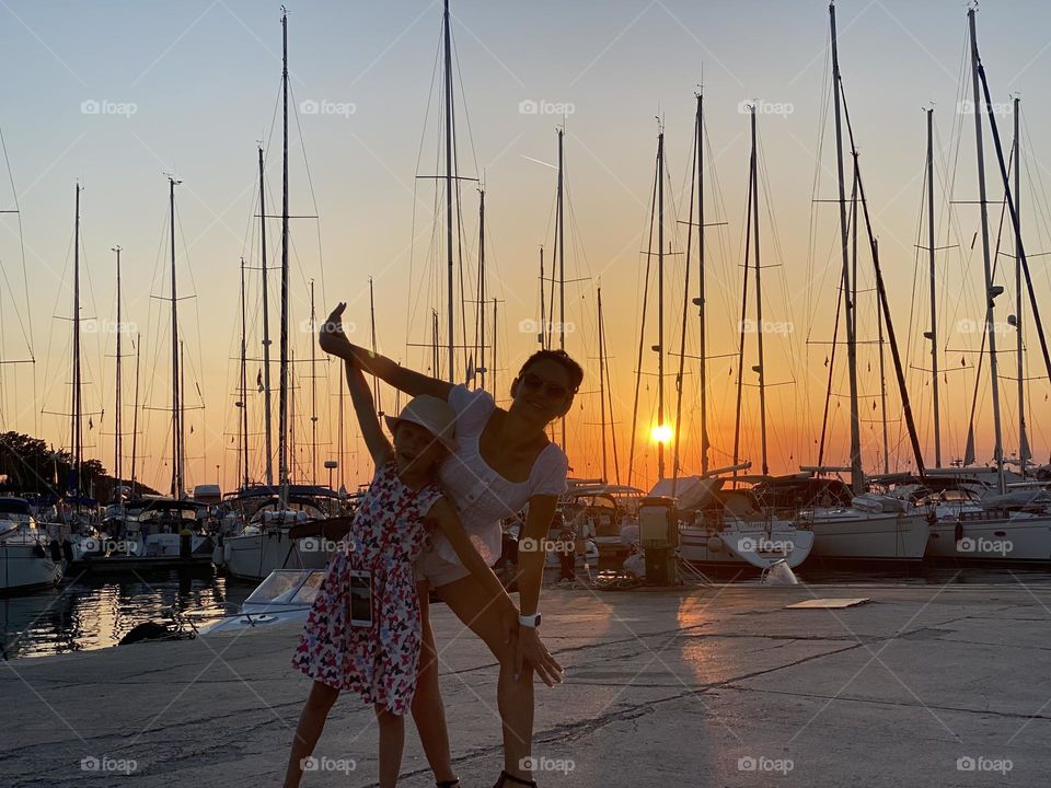 against the backdrop of a beautiful, red, summer sunset on the pier, mother and daughter against the backdrop of boats, yachts, ships