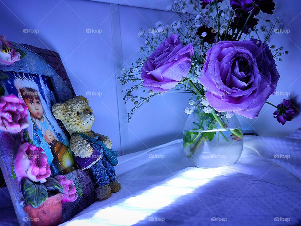 On the shelf is a jar of delicate purple roses, white small flowers and dark purple asters. Nearby is a frame with a photo of a little girl. The ceramic frame is decorated with purple flowers and a bear in a purple vest. The shelf is illuminated.