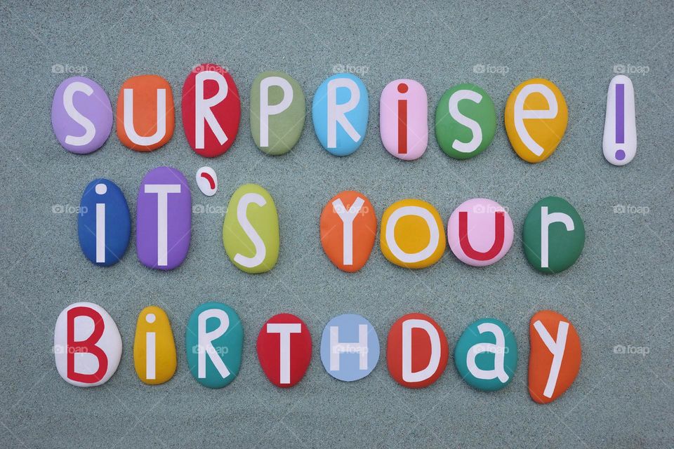 Surprise, it's your Birthday, creative message composed with hand painted multi colored stone letters over green sand