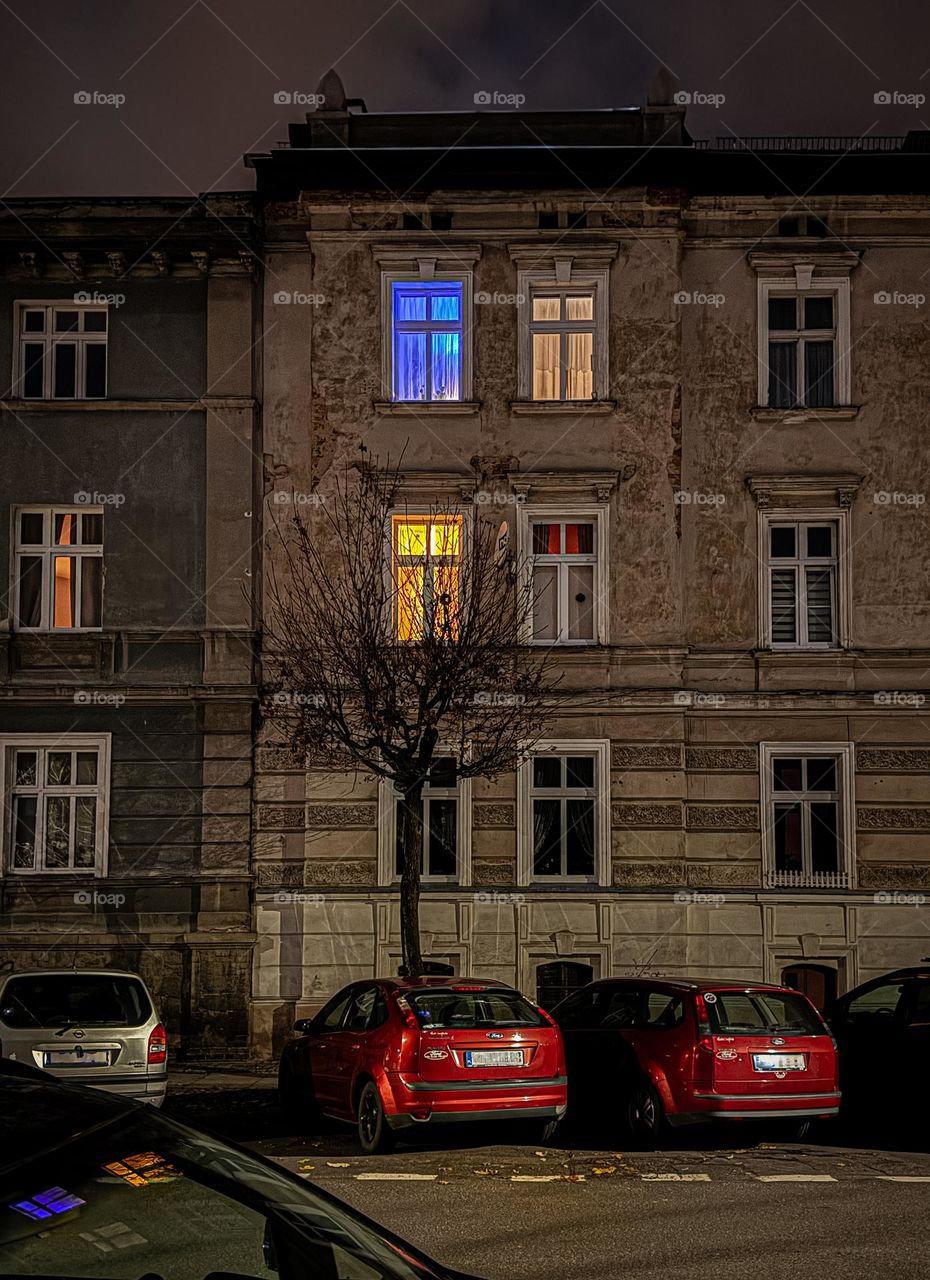 Late evening, old building, old architecture, lighting windows, blue and yellow lights, red cars on the street, reflection of the light from windows in the car window