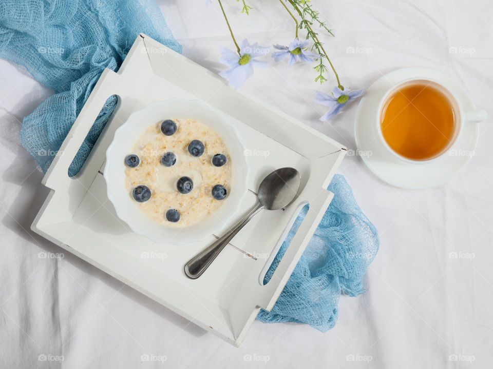 Oatmeal with fresh blueberries in a small wooden white tray with a spoon lying on a bed with a mug of tea, a gauze napkin and flowers, close-up top view. The concept of breakfast in bed.