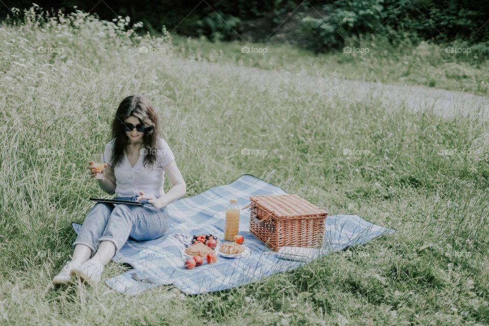 One young beautiful caucasian brunette girl with flowing hair in jeans and a t-shirt sits on a bedspread with a wicker basket, glasses, browsing social networks on a tablet while relaxing in a park on a summer day, side view close-up. Picnic time con