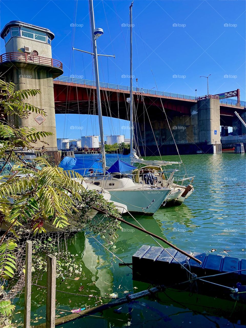 This is “Newtown Creek” and the “Pulaski Bridge” seen through the foliage of bushes on the LIC shore as well as two sailboats that are moored here. 2023. Hypnotic Productions