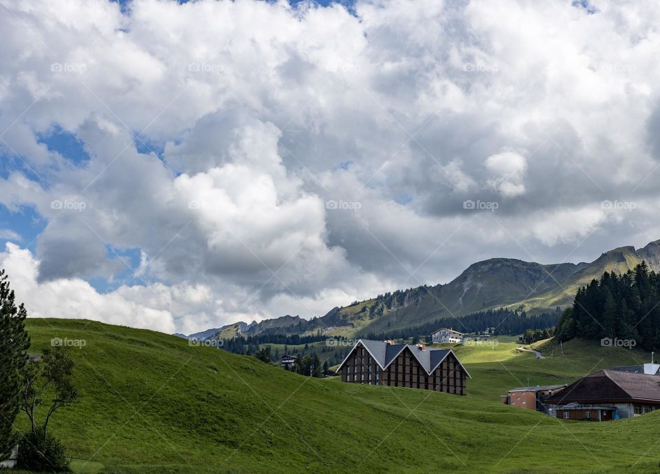 Large white and airy clouds all over the sky in the mountains with green grass, trees and a settlement with traditional houses in Switzerland, close-up side view.