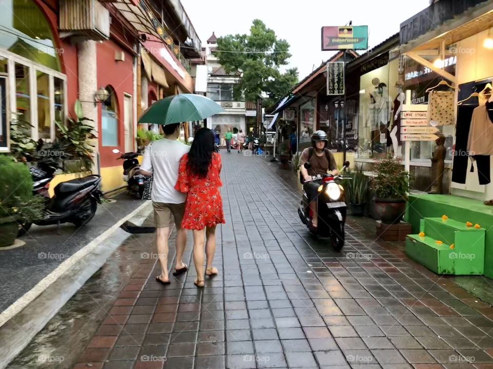 Walking in stride 5. Adult couple with open umbrella. Brick paved street. Shops. Motorcycle. Ubud, BalI. 2018.