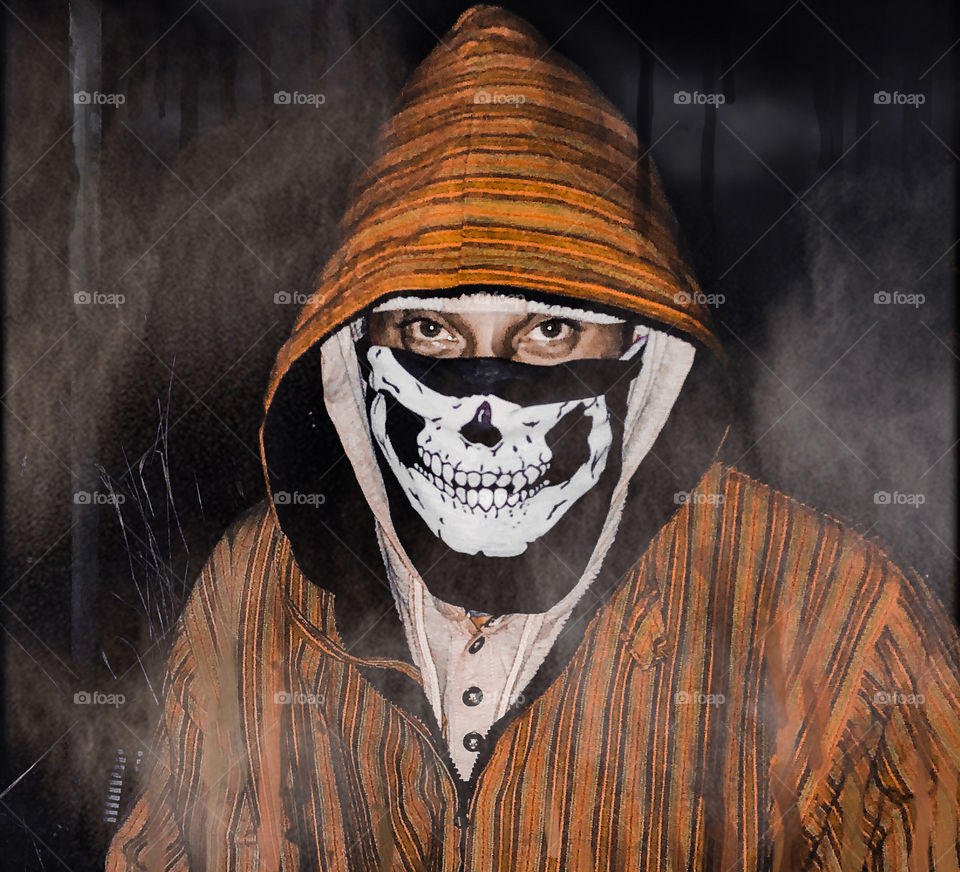 A man in an orange hoody and skeletal face mask in dark, misty surroundings