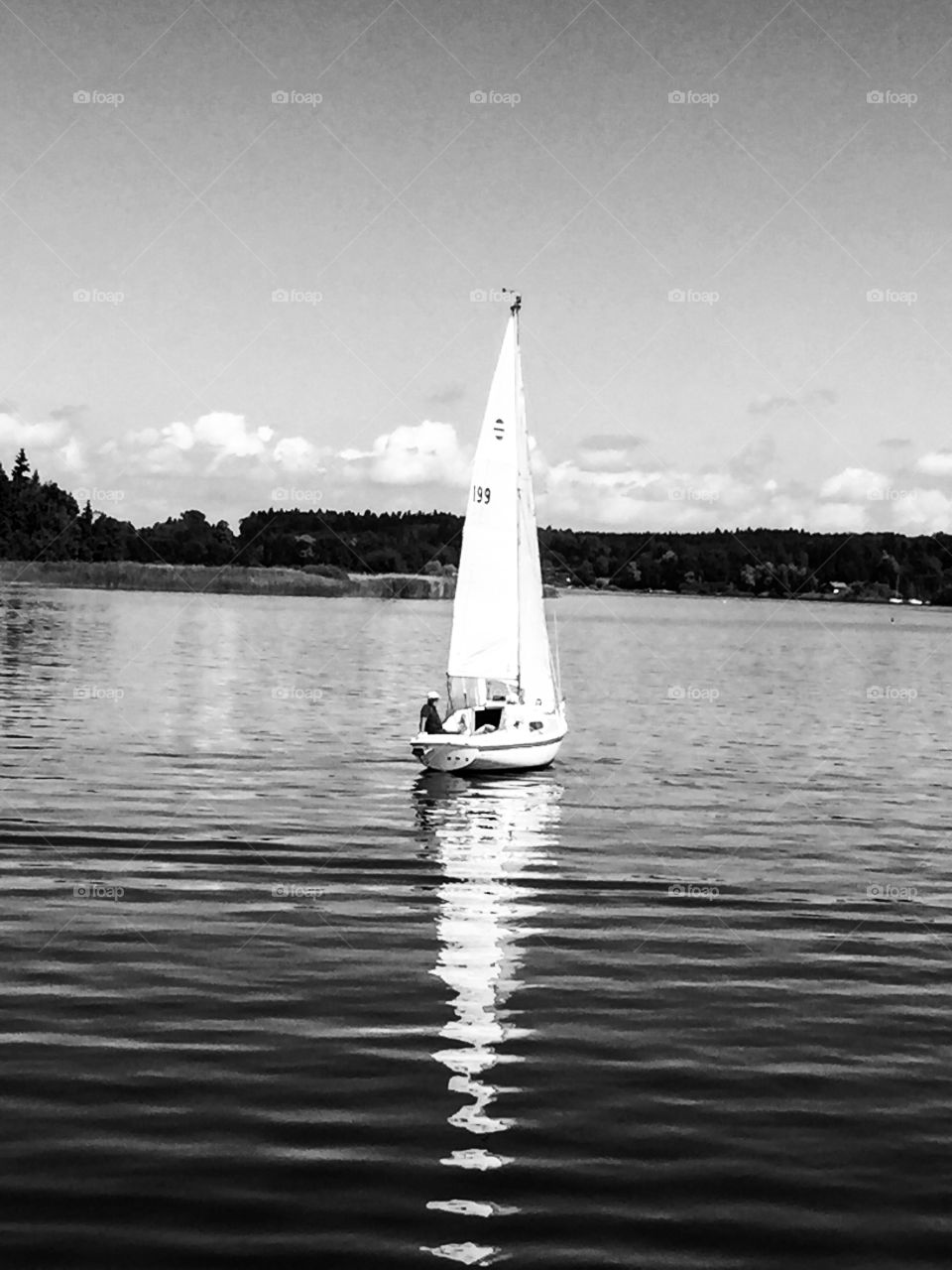 A sailboat on a lake . reflection into the water . rows of trees and clouds . black and white image. man is relaxing on a sailboat