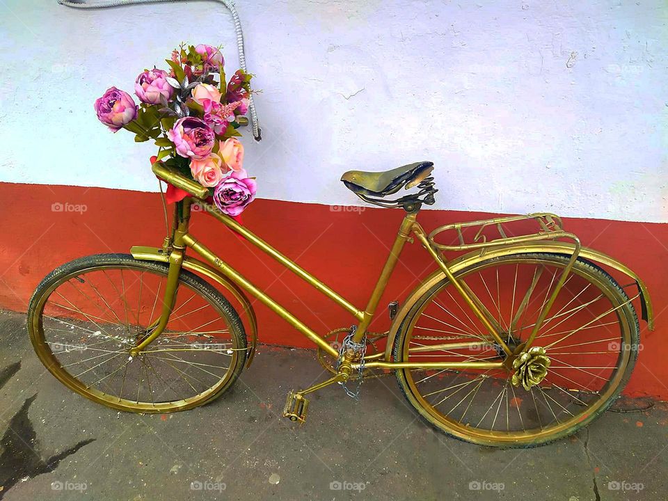 Bicycles. Near the wall is a bicycle painted in golden color. Flowers attached to the front frame of the bicycle