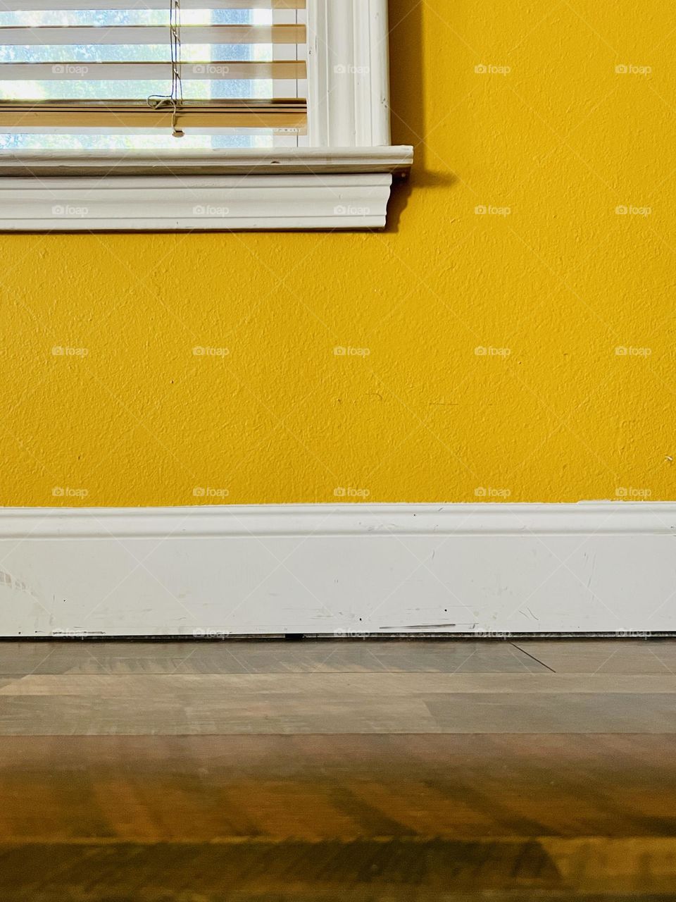 Closeup interior wall painted deep yellow, with white baseboard and window frame, and a wooden laminate flooring 