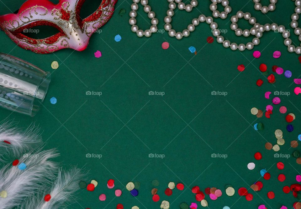 Masquerade mask, champagne glass, white pearl necklace lie framed on a green background with copy space in the center, flat lay close-up. The concept of carnival, masquerade, holiday banner.