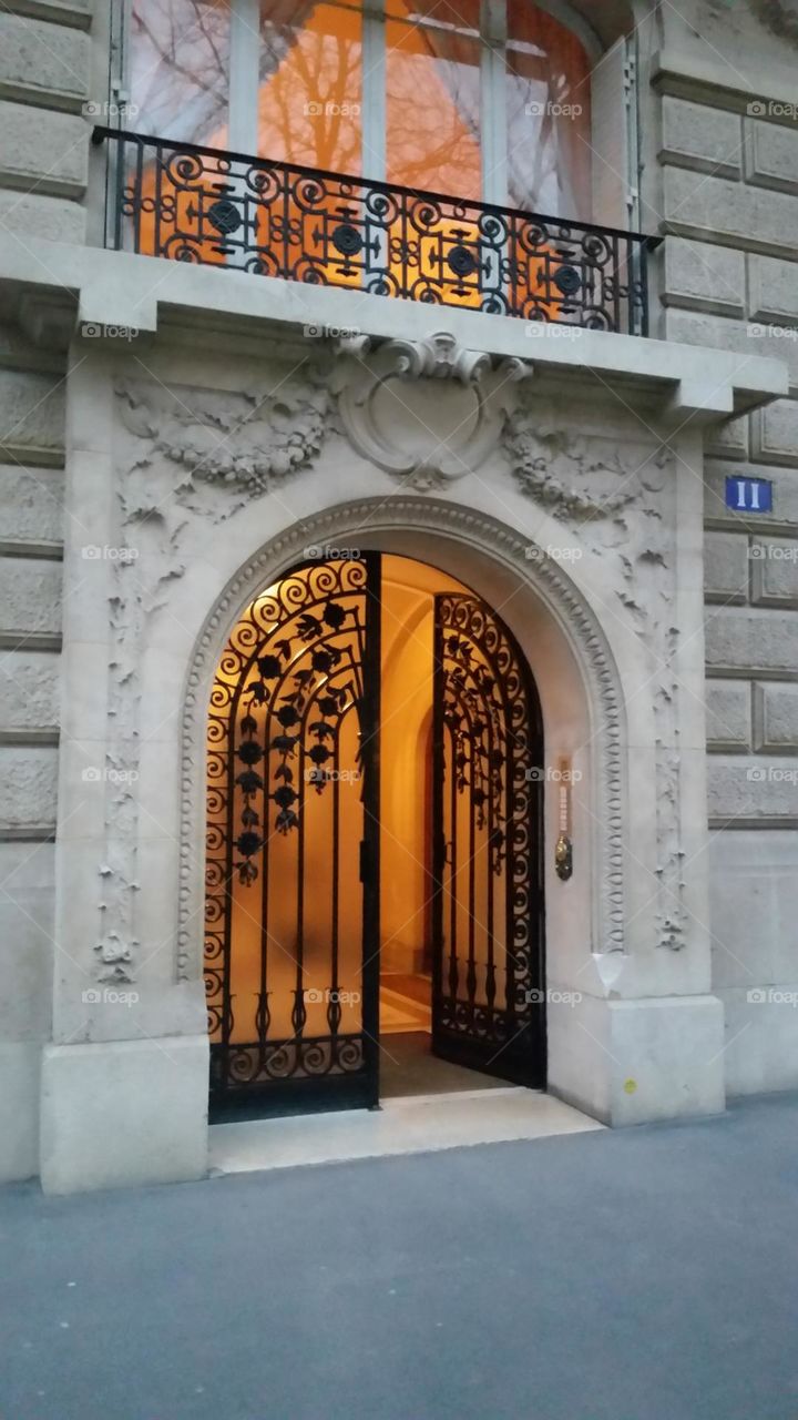 Illuminated paned window with cast iron balcony rail and cast iron entrance door of a stone-built Haussmanian building in Paris at nightfall