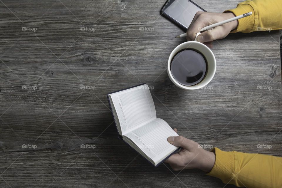 A man drinks coffee in the morning, works on the phone and makes notes in pencil in a notebook. Men's hands hold a white cup of coffee and a mobile phone in a yellow jacket. The hands lie on a gray wood background.