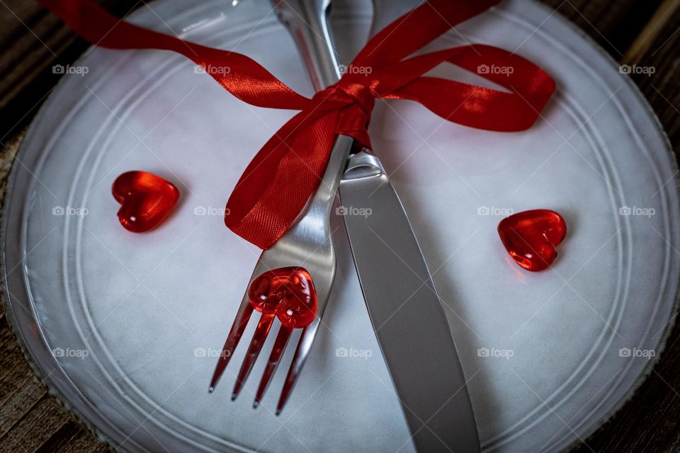 A beautiful view of a fork and knife tied with a red ribbon and a glass heart lying on a white empty plate on a brown wooden table, side view.