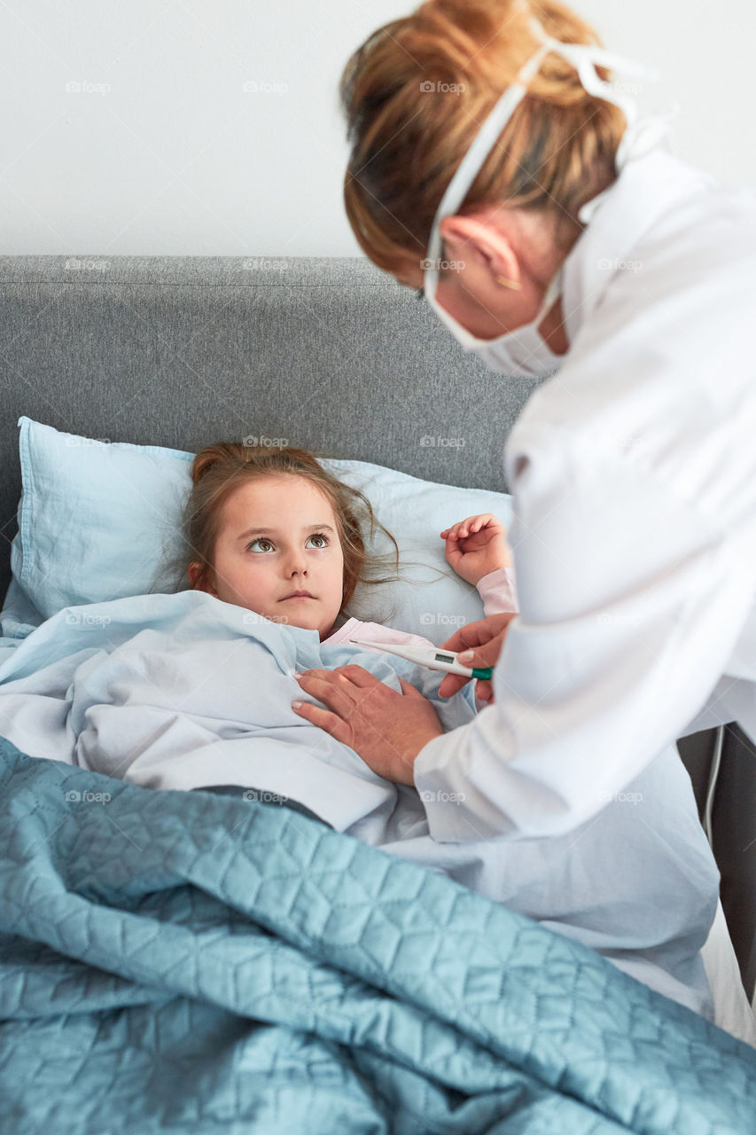 Doctor visiting little patient at home. Measuring the temperature of sick girl lying in bed. Woman wearing uniform and face mask. Medical treatment