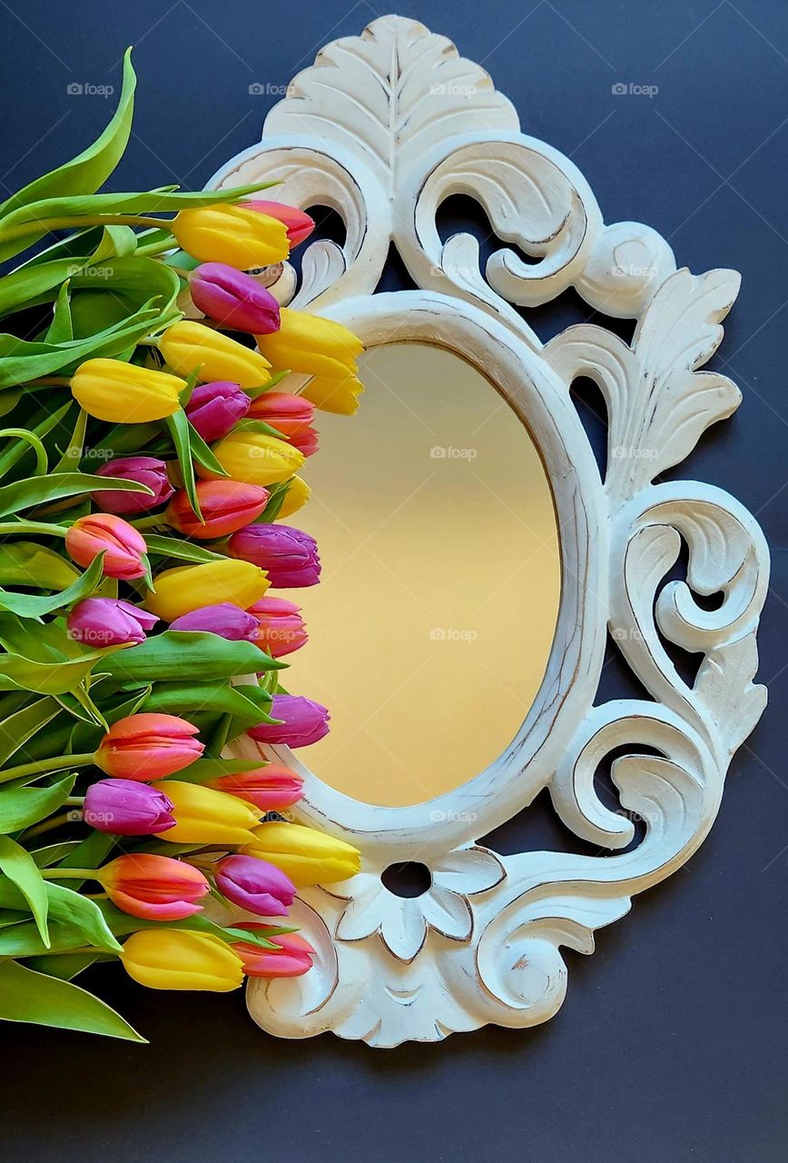 Spring primary colours flatlay on a black surface made of a carved wood mirror and a bunch of yellow and purple tulips spread along the left side of the mirror