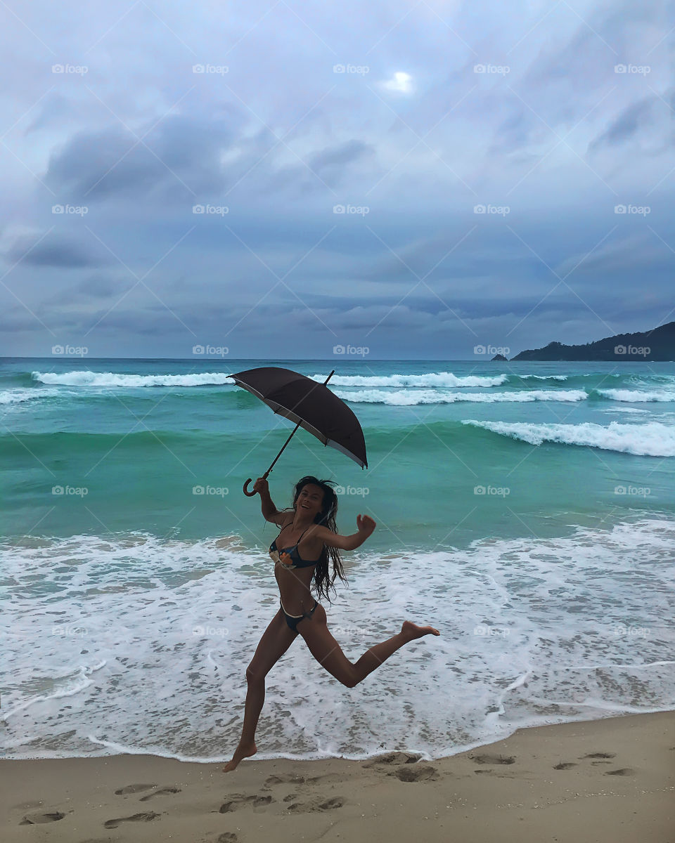 Young woman enjoying the tropical rain under umbrella at the sandy beach 
