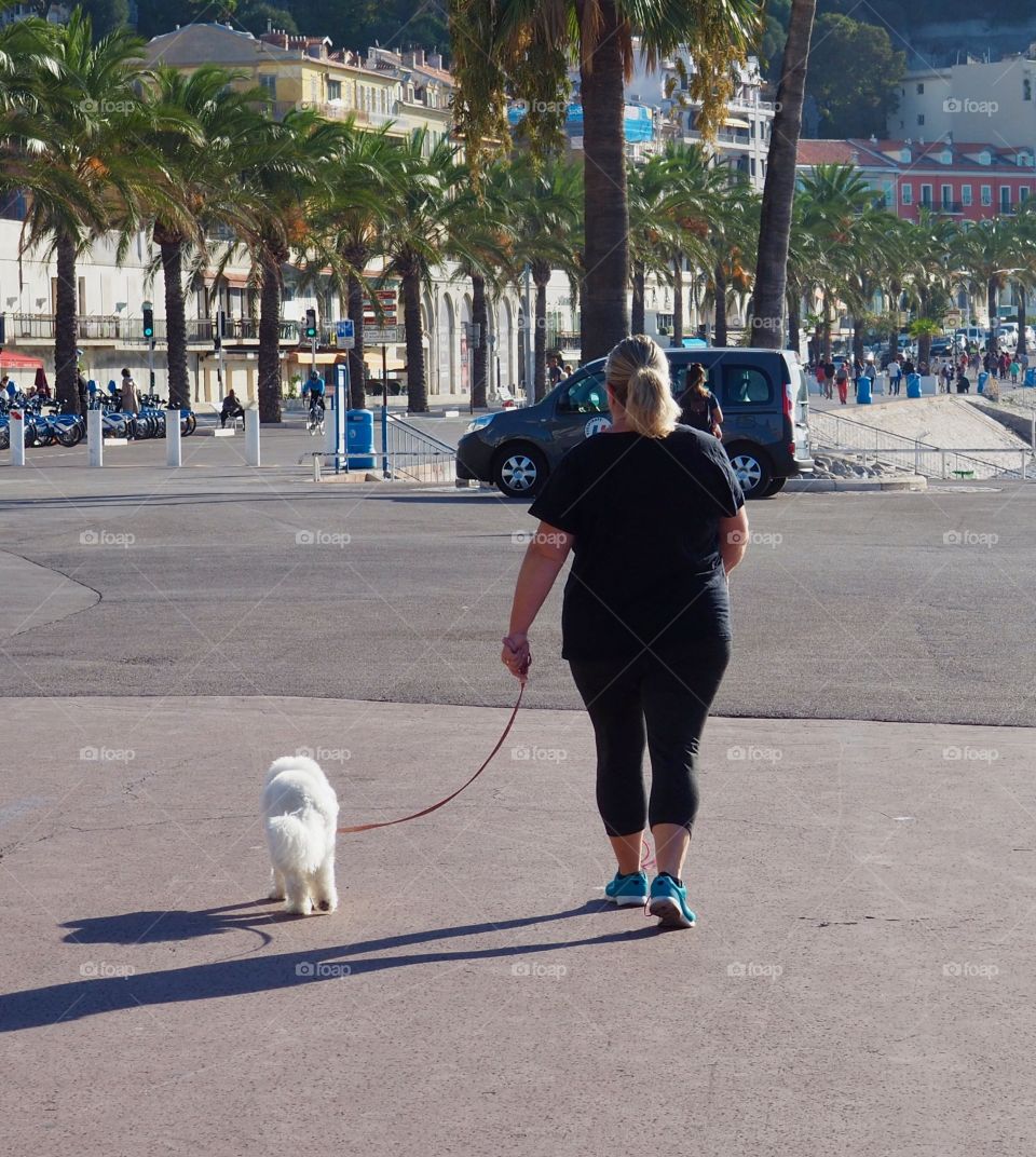 Overweight woman in workout clothes walking with her white small dog on the Promenade des Anglais.