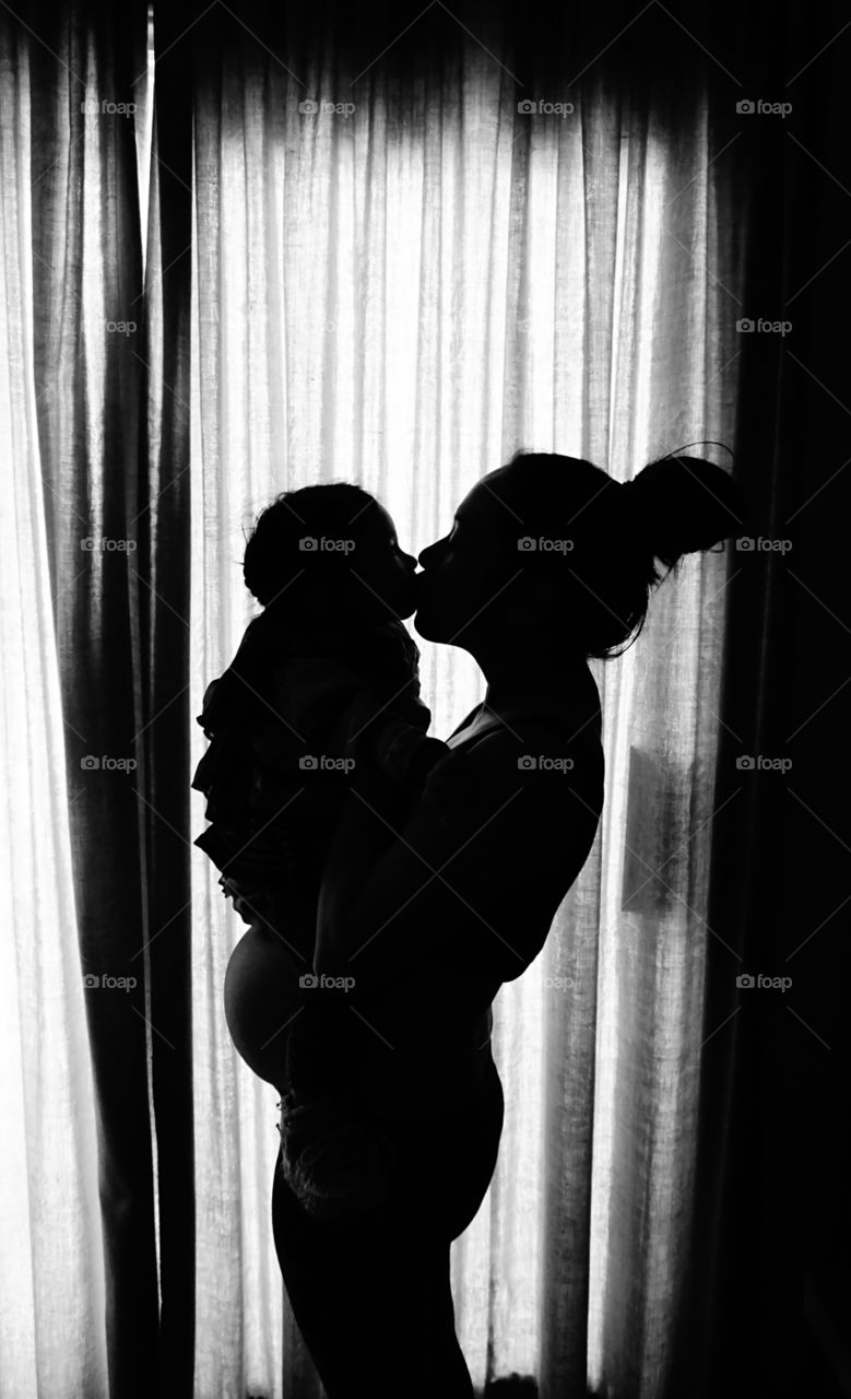 Silhouette of a mother carrying her daughter on her pregnant belly and giving her a kiss.