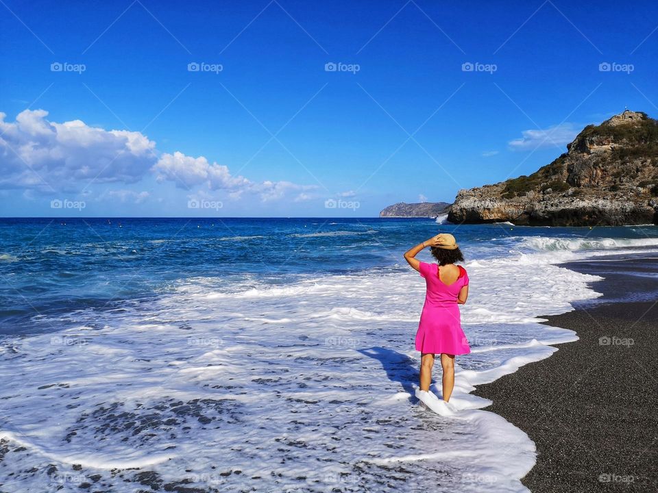 seen from behind, woman in fuchsia dress wets her feet by the sea