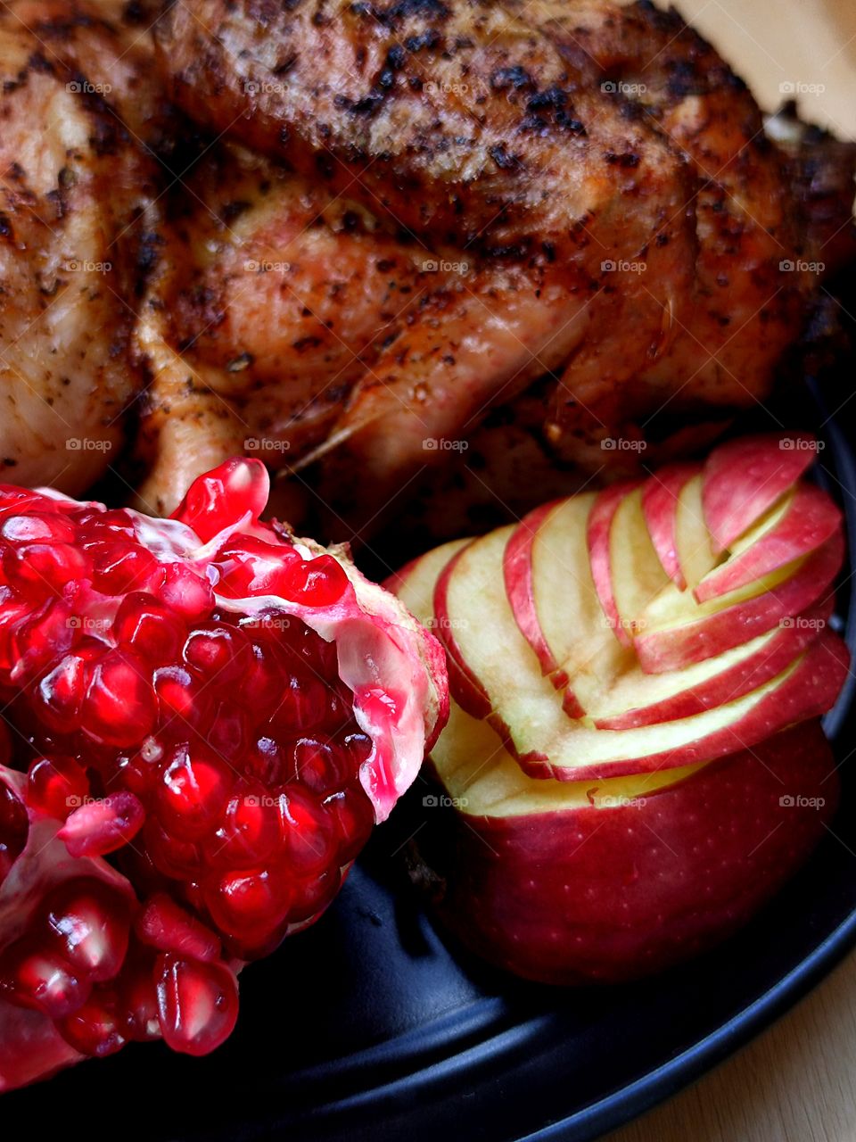 Composition.  On a black platter are: half a grilled chicken, a cut pomegranate and a red apple in the form of a leaf