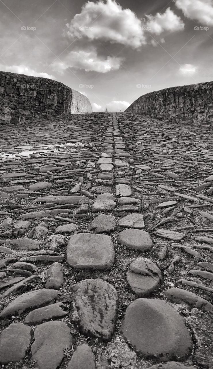 A view from the ground going up over a cobblestone arched bridge reaching up to the sky 