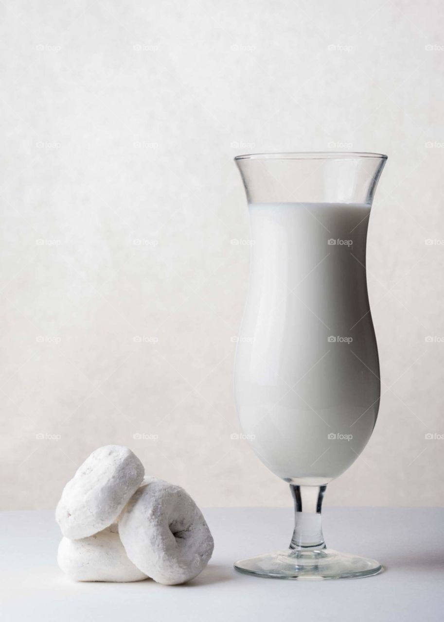 white milk in a glass next to mini white powder doughnuts on a white table in front of a white background
