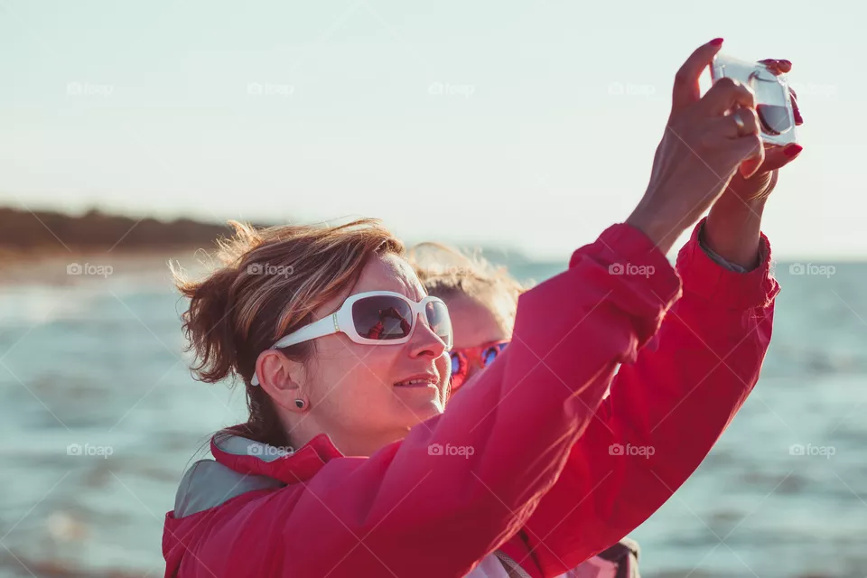 Young woman taking a photos using phone, looking at screen, standing outdoors, she is backlighted by sunlight with plain sky and sea in the background