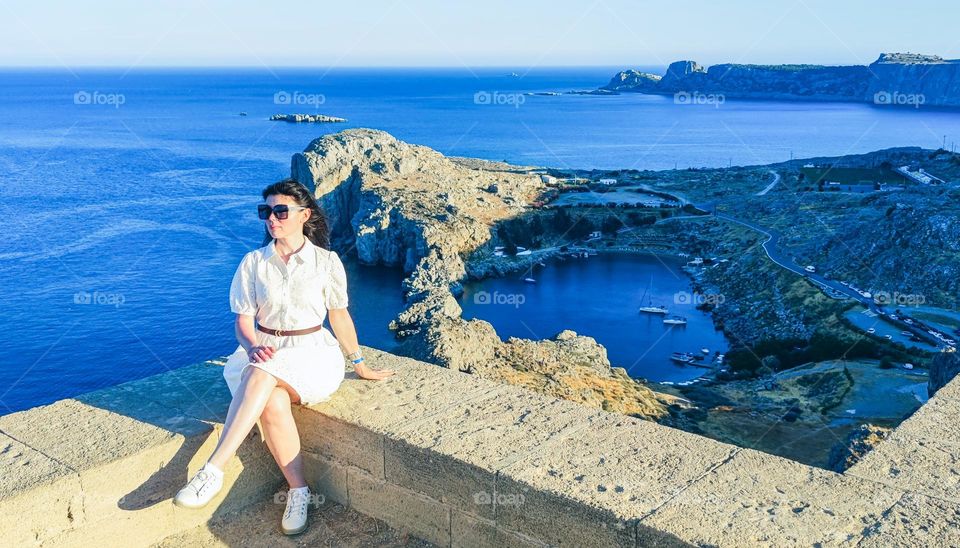 A beautiful young Caucasian travel girl in a white openwork dress, sunglasses and sneakers sits on a stone curb of an ancient temple to the goddess Athena looking to the side against the backdrop of the Mediterranean sea with the bay of St. Paul