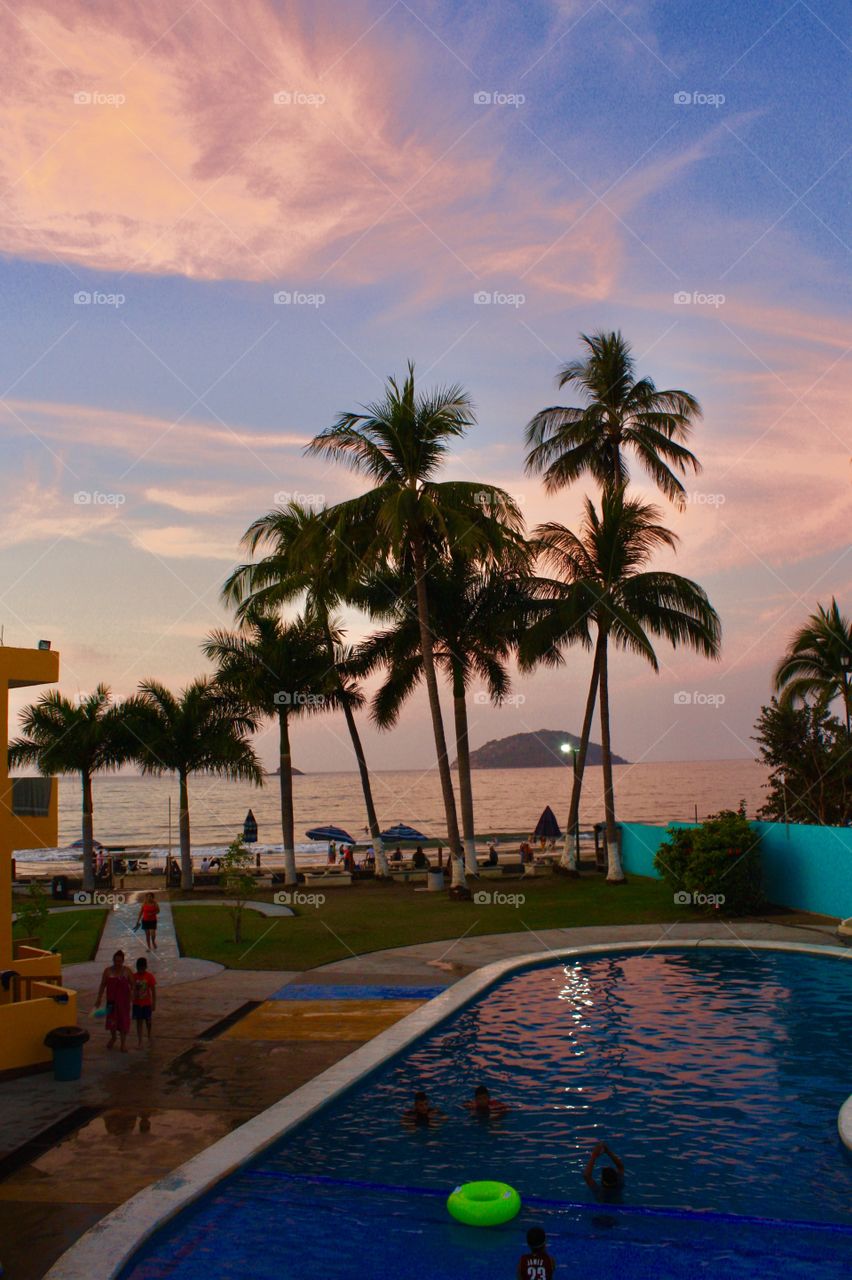 Exterior daylight.  Guyabitos Beach, la Peñita de Jaltemba, Mexico.  Sunset on a hotel’s pool, palm trees, the sea, and Isla de Coral.