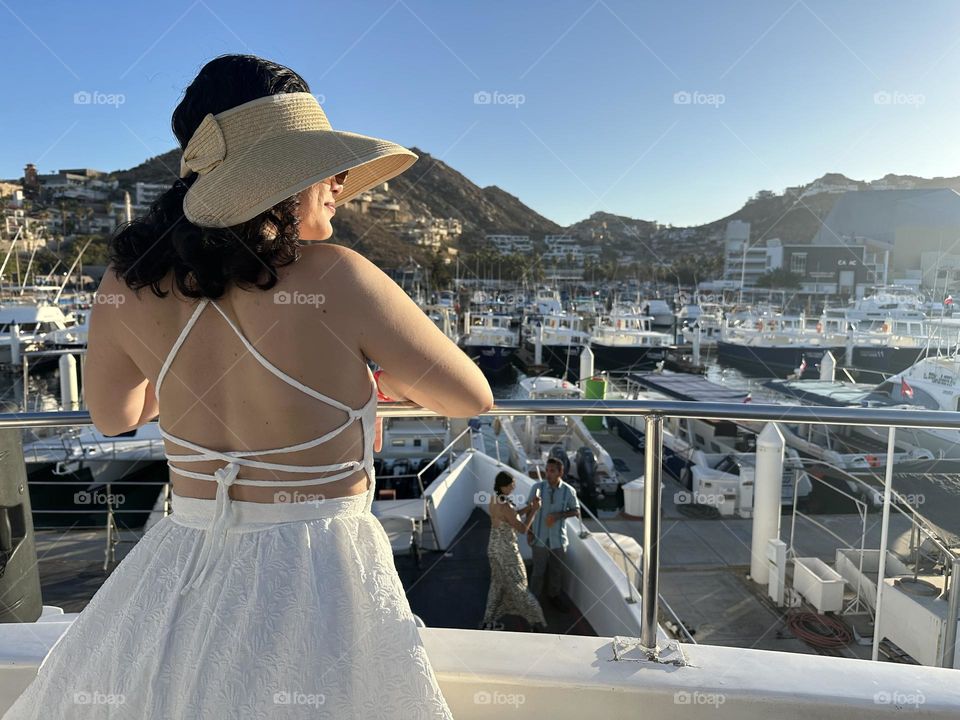 Woman wearing a hat, standing on a boat, looking at the horizon on a beautiful sunset.