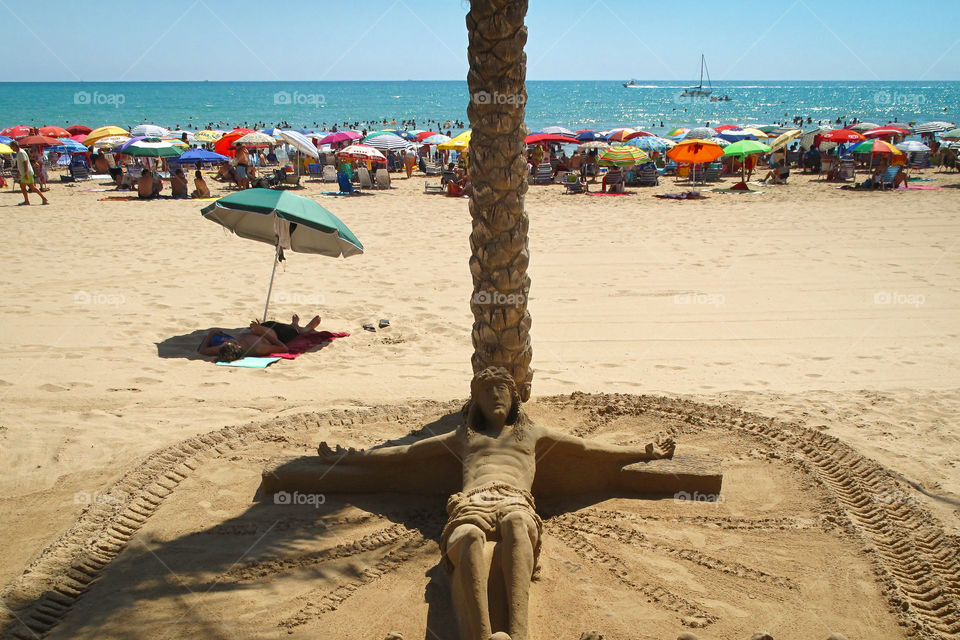 Jesus Christ on vacations. Sand sculpture depicting crucified Jesus lying down under palm tree.