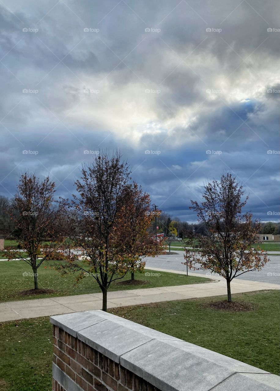 Big gray clouds illuminated by the setting sun over a parking lot in Midwest Michigan that has fall trees and green grass surrounding it