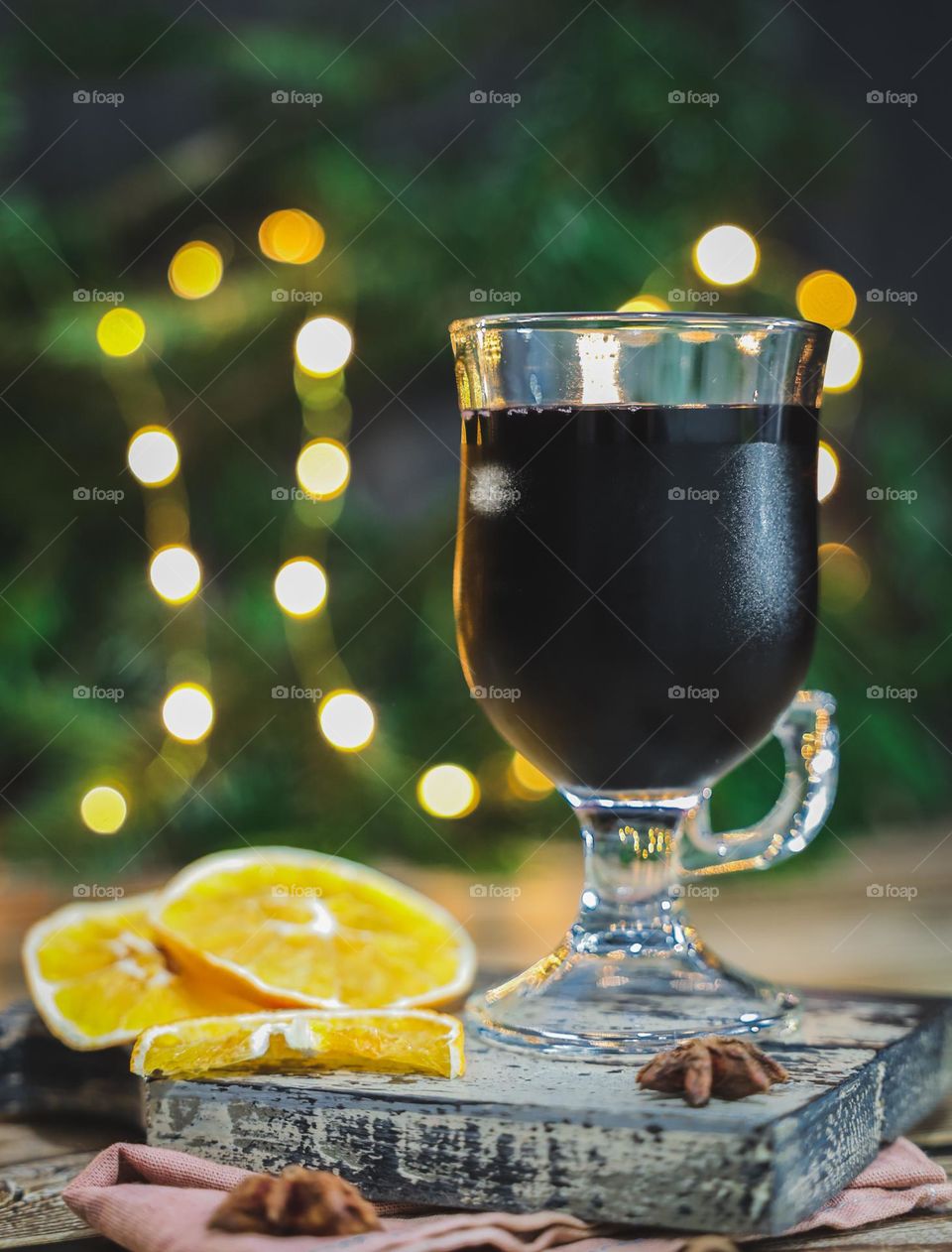 One glass of hot red mulled wine with anise and orange slices on a cutting board stands on a wooden table on a blurred background with a Christmas tree, a garland and bokeh, close-up side view. Holiday traditional drinks concept.