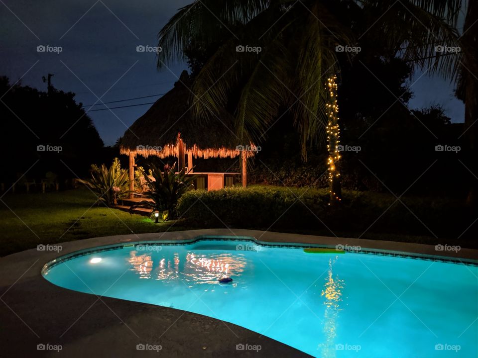 Nighttime view of a lighted dugout swimming pool with palm trees and tiki hut in the background