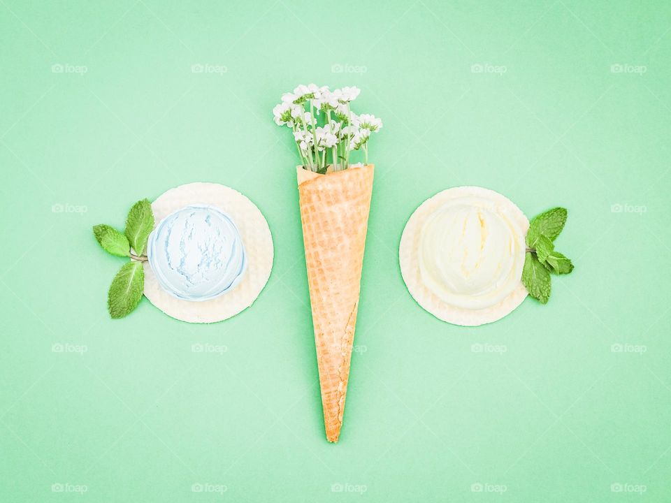 A waffle cone with small white gerberas and pastel-colored popsicles with mint leaves on round waffles lies on a light green background, a close-up flat lay.