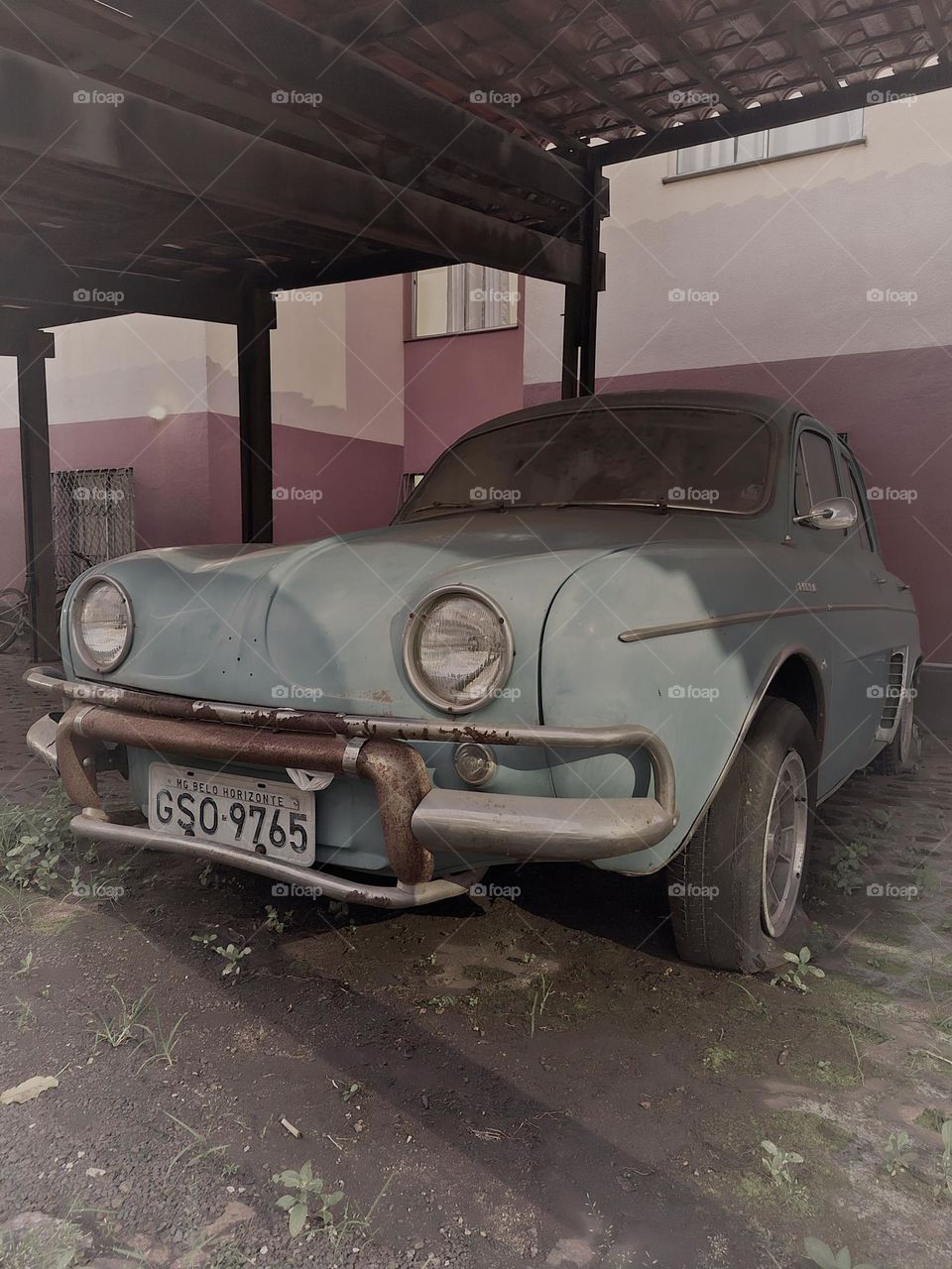 A classic light blue Renault Dauphine, showcasing its vintage charm with signs of wear, parked under a roofed structure. The aged paint and rusted bumper details give the car a nostalgic character, ideal for automotive relic enthusiasts.