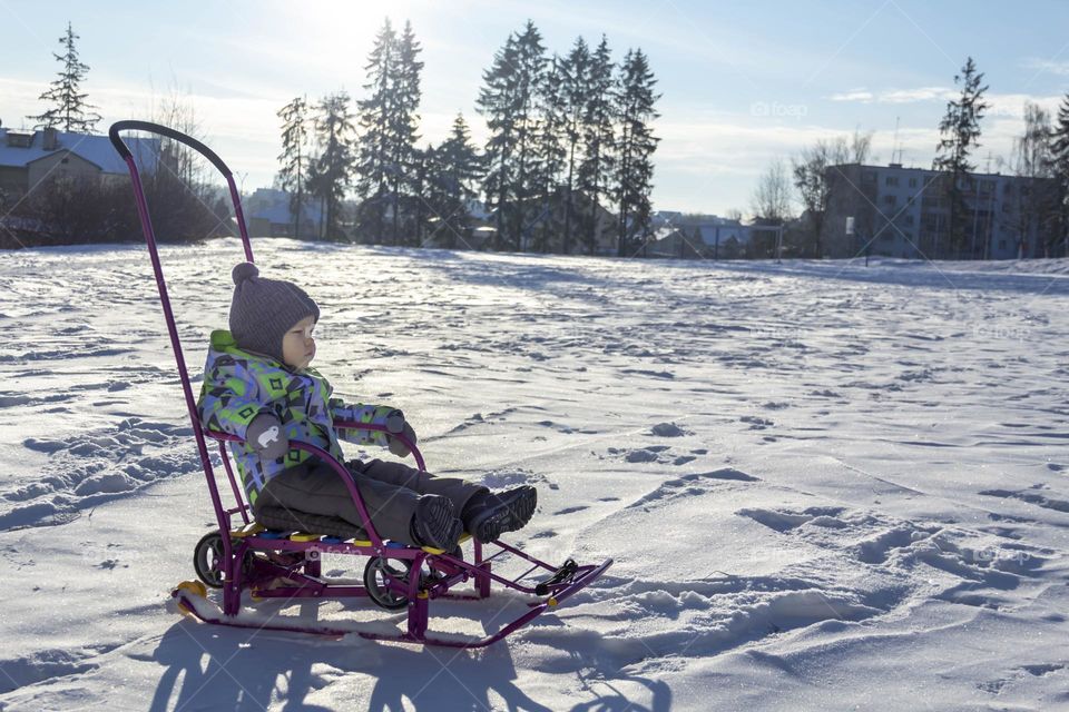 A child in winter in winter jackets, pants, a hat and boots on white snow on the street and in the park in nature plays winter fun and sleds.