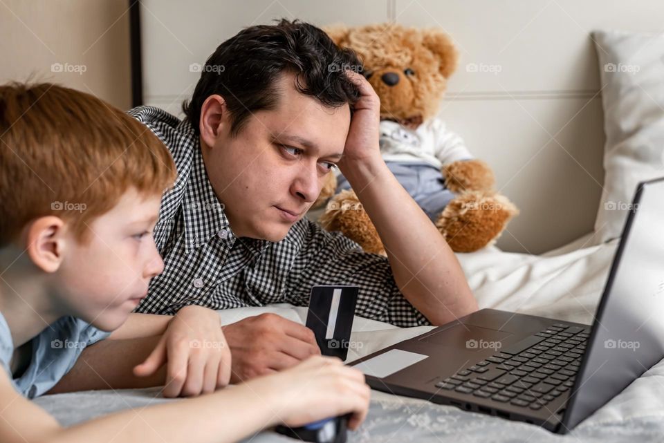 Father and son have fun together, at home they buy toys online shopping sit by the bed with a laptop, close-up portrait life style