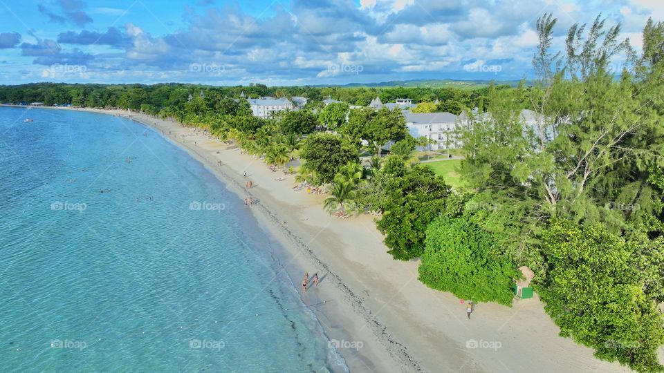 From above, Seven Mile Beach in Negril, Jamaica is a breathtaking spectacle. Turquoise waters meet a ribbon of white sand, while emerald palms sway on the fringe. Tiny figures - visitors, enjoy this piece of paradise under the radiant Caribbean sun.