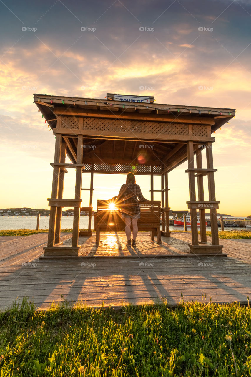 Beautiful young woman watching the sun setting over an island, standing in a cute wooden gazebo. 