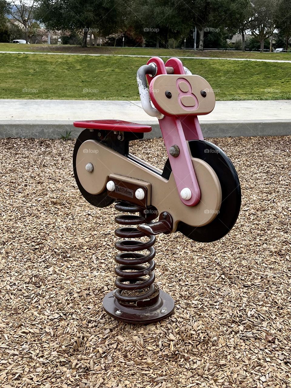 A cute red Children’s bouncy bike ride in a public park.  It’s sitting in the tan bark from the playground with the sidewalk and a green grassy field behind it.  Fun for kids