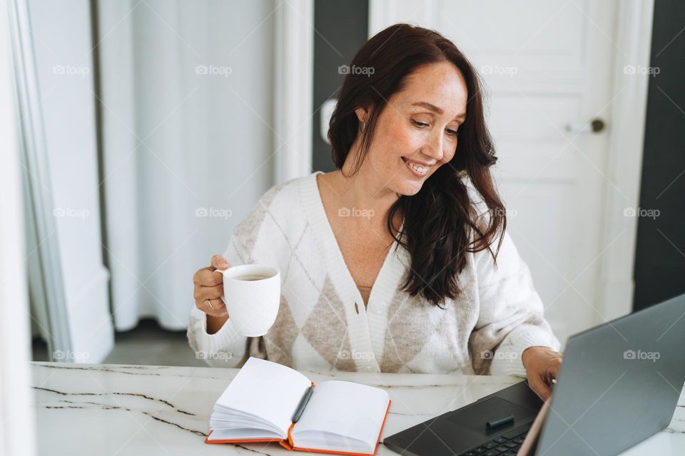 Smiling brunette woman with long hair in white cardigan working on laptop in bright modern office