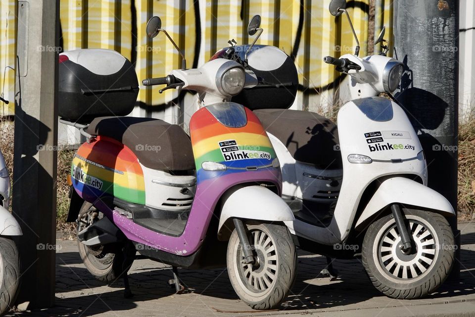 Two blinkee.city mopeds, which can be driven with a B driving license, are parked on a sidewalk in Prague-Palmovka. One moped is in the colors of the rainbow flag.